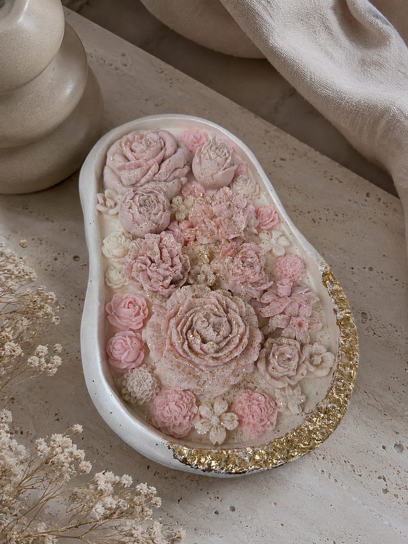 Große handgefertigte Blumenkerze in weißer Gipsschale mit rosa Blüten und goldenen Details, gehalten in der Hand.